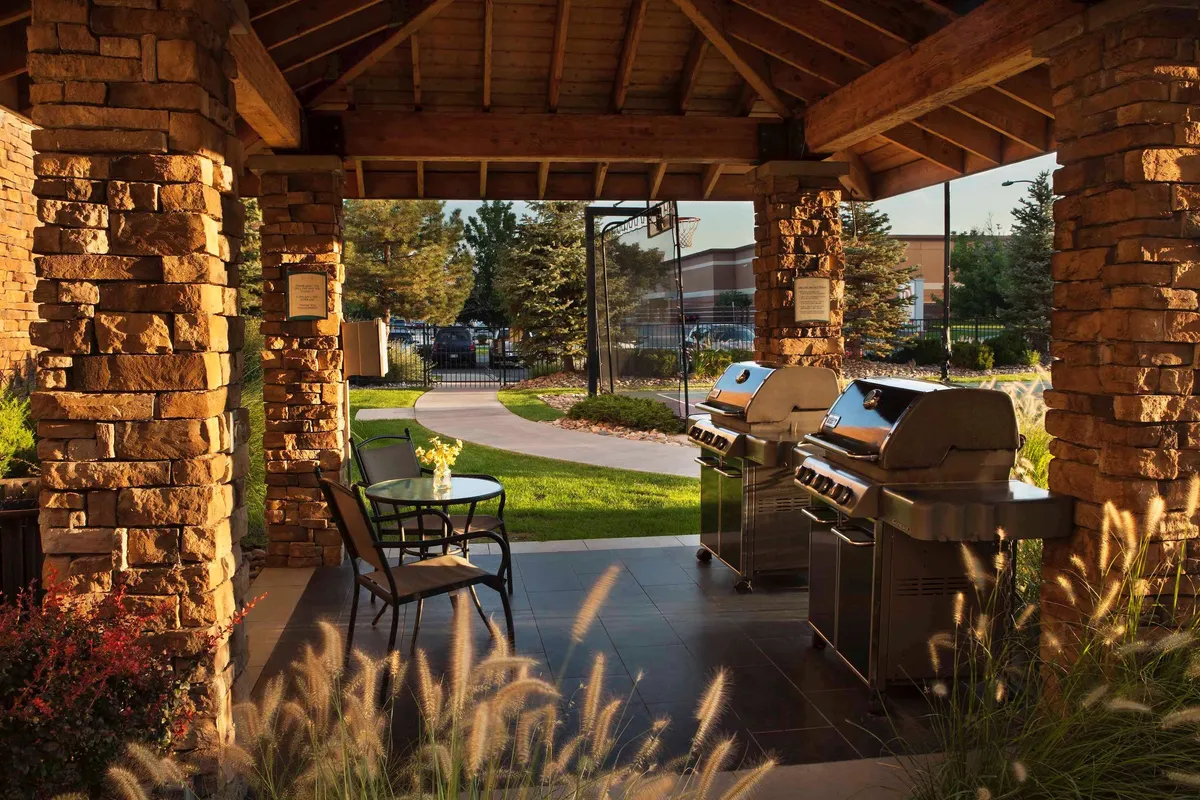 This exterior image showcases inviting grill stations and an outdoor seating area nestled within a beautifully landscaped garden. The scene is set during sunset, capturing a group of people enjoying a relaxed barbecue, with lush greenery and natural surroundings emphasizing the outdoor lifestyle.