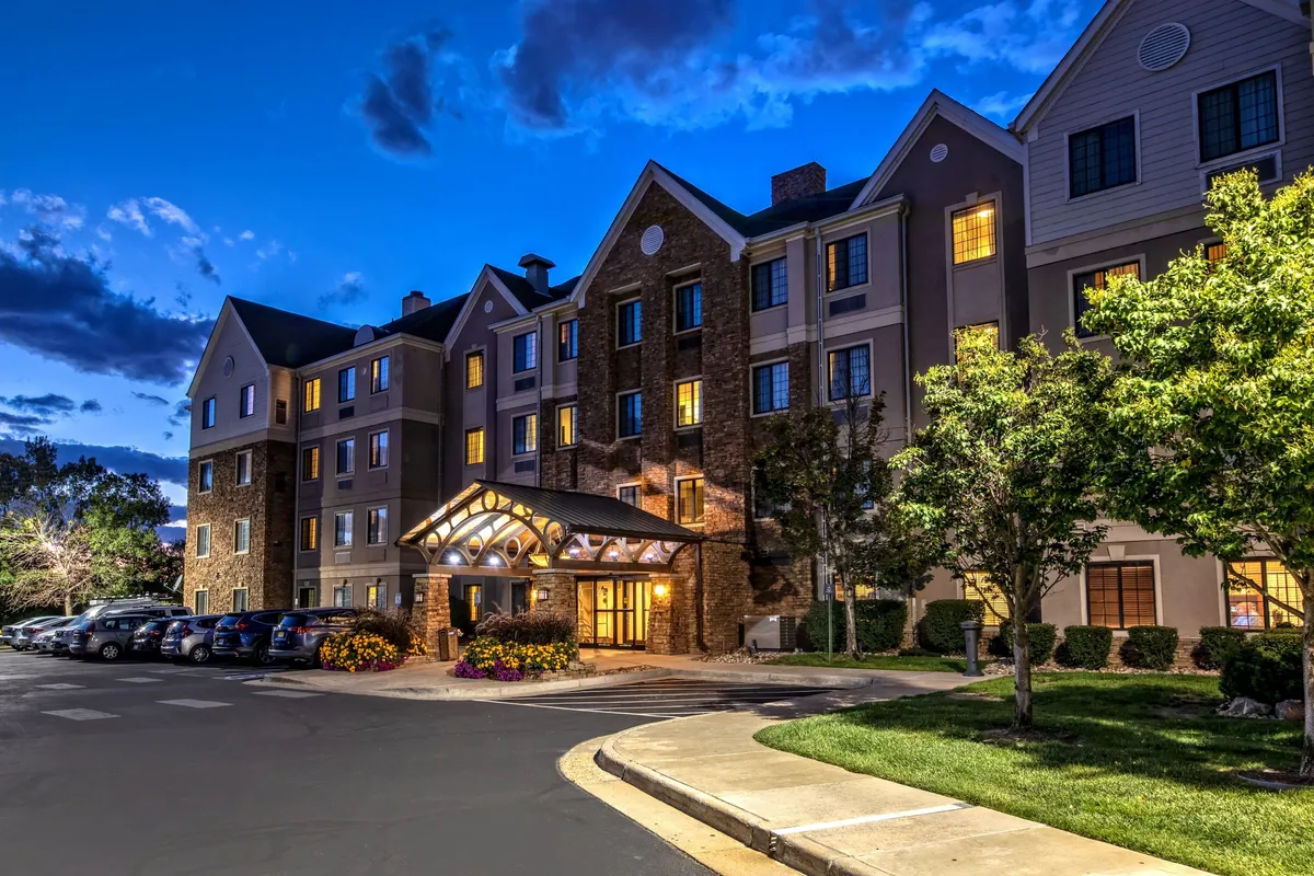 The exterior of the building is shown in the evening, with a welcoming entrance awning, softly lit windows, and well-maintained landscaping. Several cars are visible, highlighting the on-site parking area. The scene feels inviting and serene, suggesting ease of access and proximity to nearby attractions.
